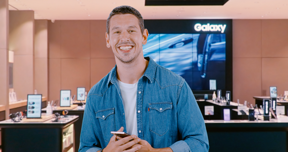 Ator João Vicente de Castro sorrindo para a câmera no interior de uma loja Samsung