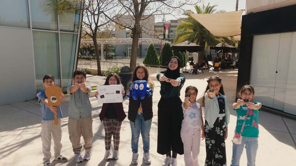 Rahaf em &aacute;rea externa junto a alunos de seu workshop, mostrando os projetos que criaram durante as aulas