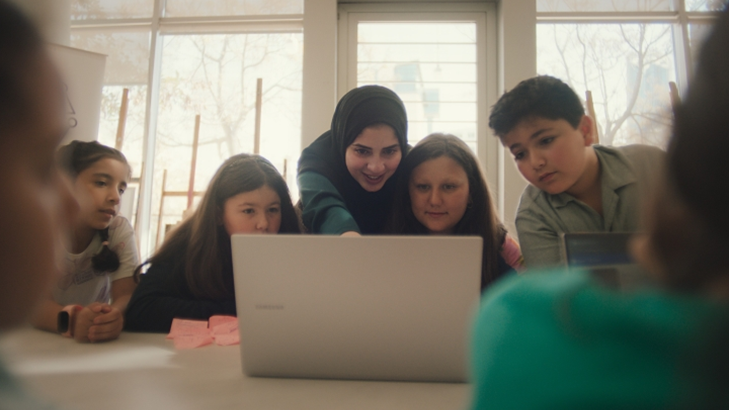 Rahaf cercada de crian&ccedil;as em uma mesa apontando para a tela de um notebook Samsung durante um de seus workshops